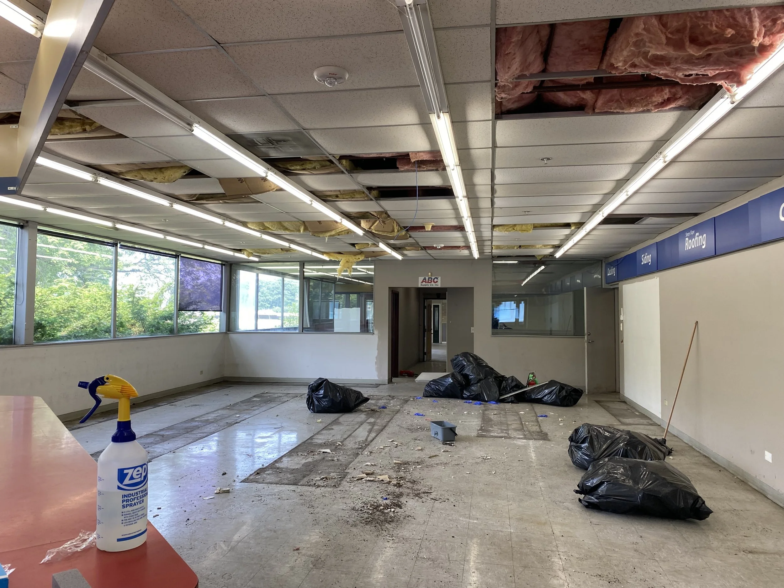 Empty shopping mall or retail space undergoing renovation, with damaged ceiling tiles, exposed insulation, scattered garbage bags, and cleaning supplies. Warehouse cleaning