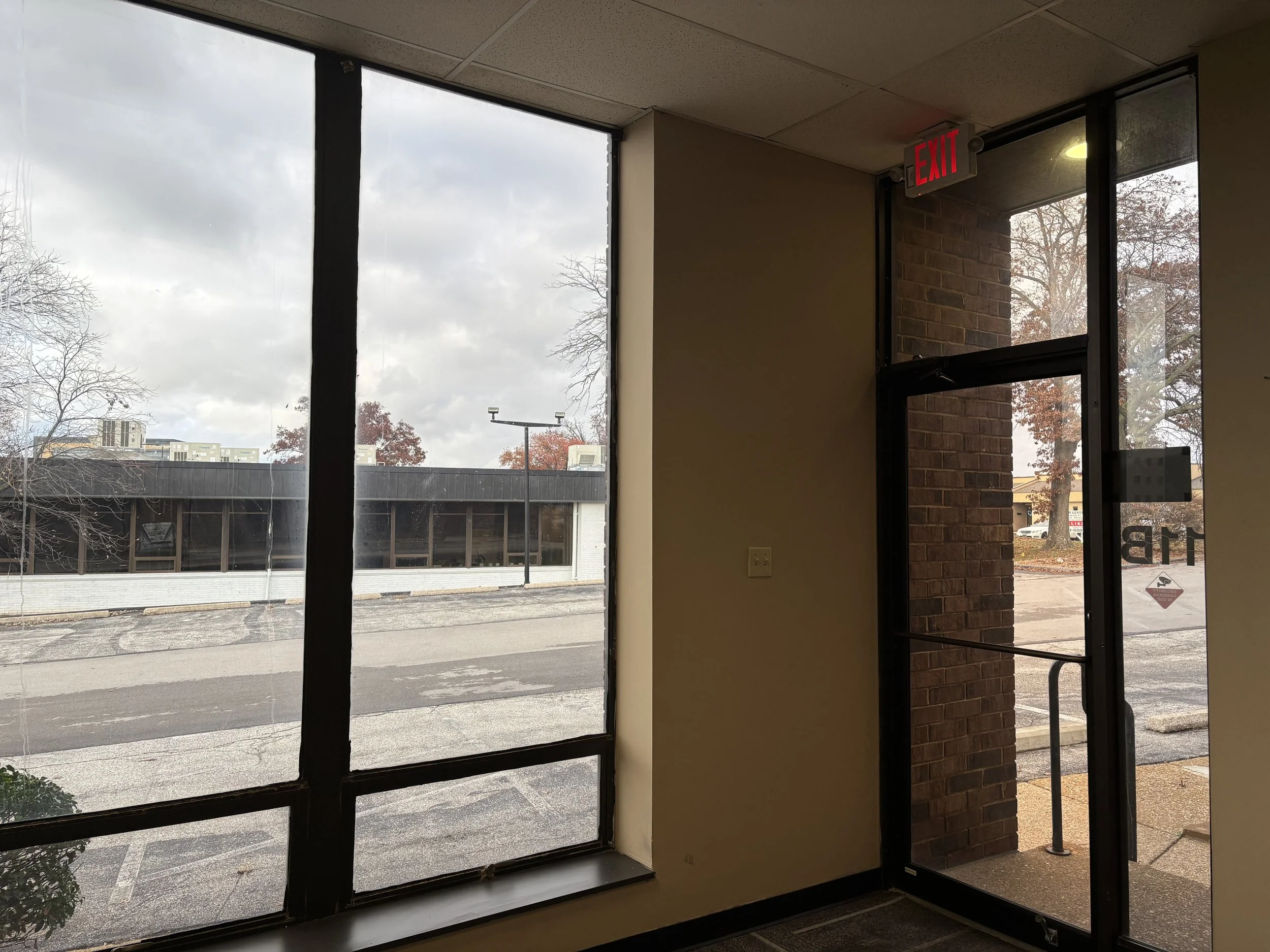 Interior view of a building entrance with a large window and glass door, showing a parking lot and street outside on a cloudy day; the door has an illuminated red exit sign above. Window cleaning