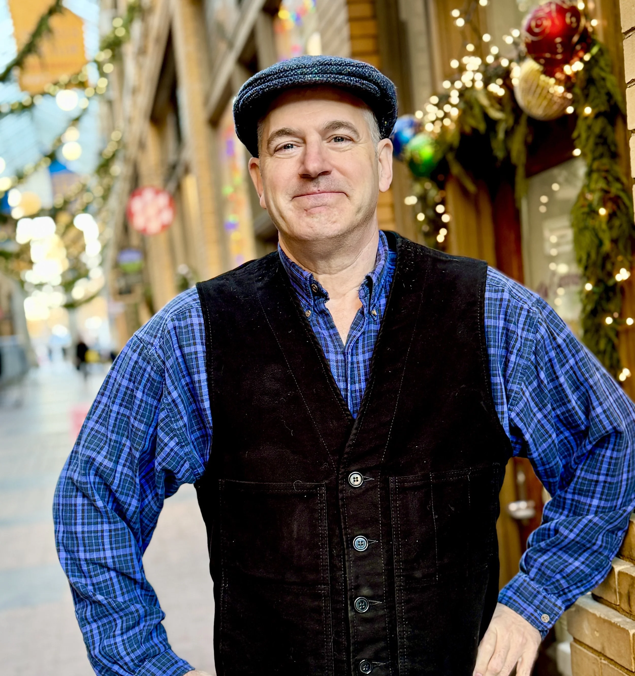 A middle-aged man standing outdoors in front of a decorated holiday display, wearing a blue plaid shirt, black vest, and a navy flat cap, smiling at the camera.