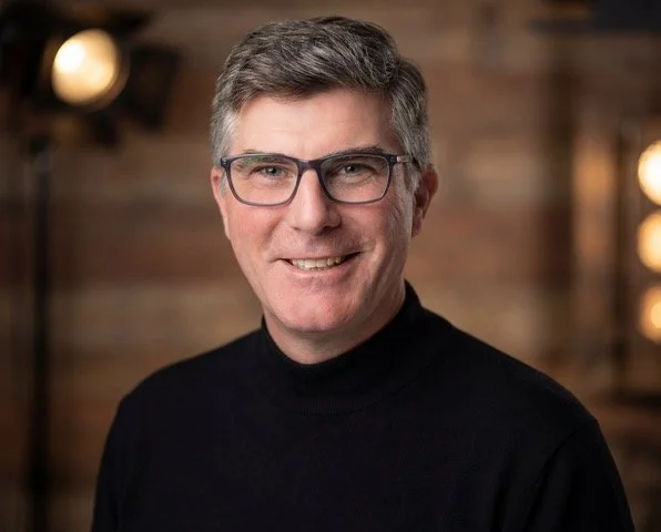 A middle-aged man with gray hair, glasses, and a black turtleneck sweater smiling in a warmly lit studio with wooden walls.