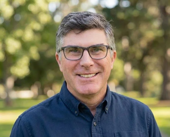A man with glasses smiling outdoors in a park, wearing a dark blue button-up shirt.