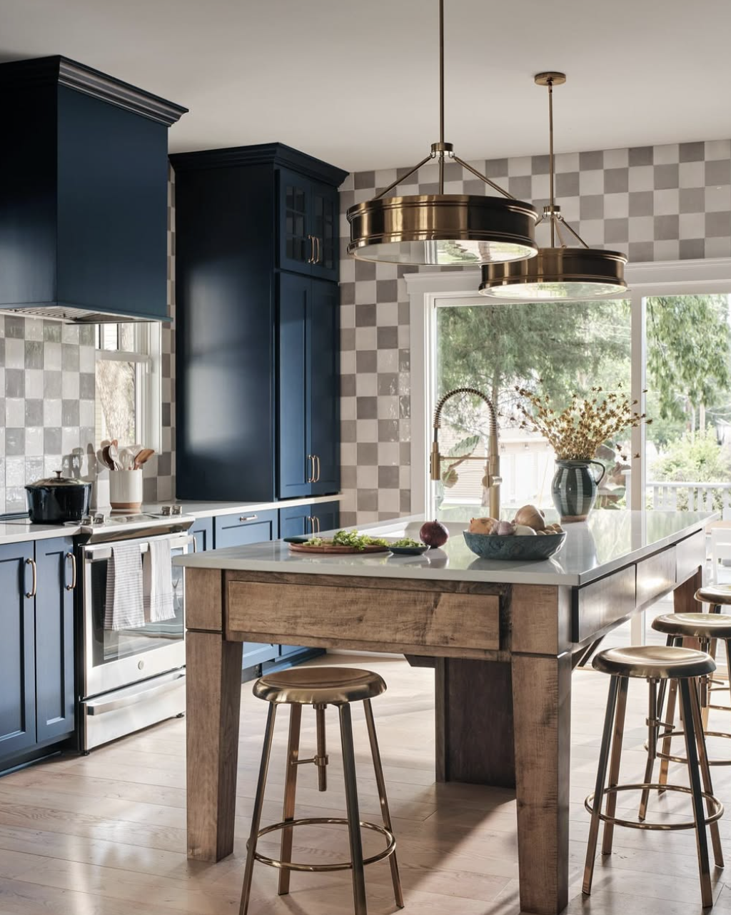Modern kitchen with blue cabinets, checkered backsplash, and a wooden island with stools, large window, and gold pendant lights.