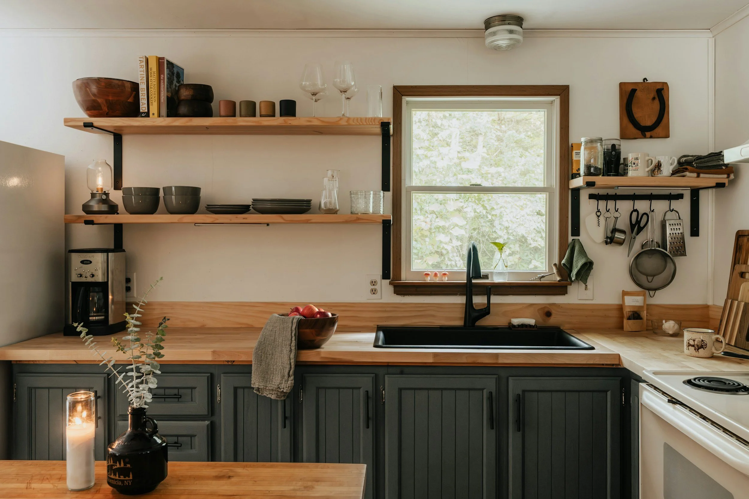 Kitchen with wooden countertops, gray cabinets, open wooden shelves holding bowls, plates, glasses, and books, a coffee maker, a window above the sink, and various kitchen utensils hanging on the wall.