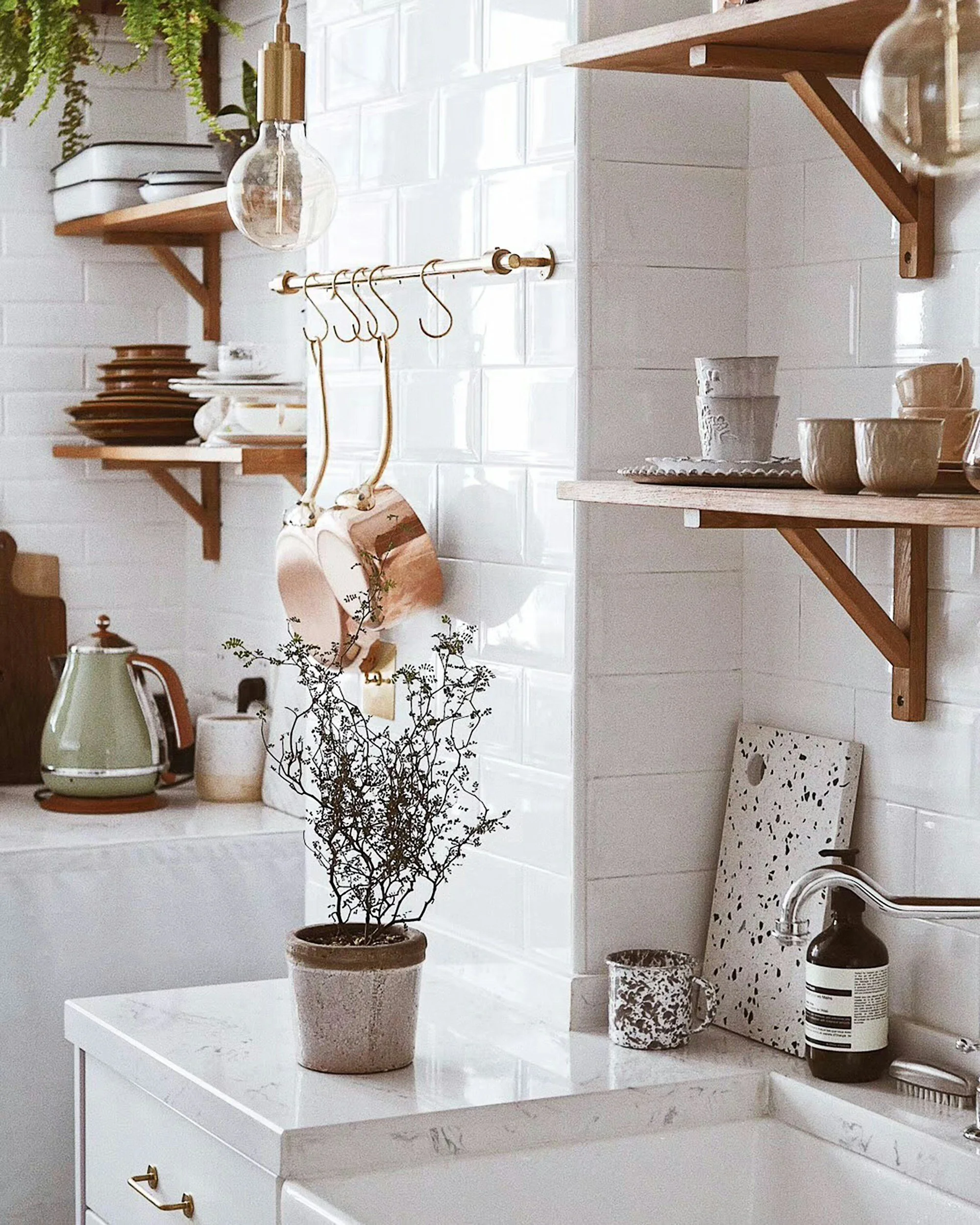 Kitchen with white tile backsplash, open wooden shelves holding dishes and cups, hanging copper mugs, a potted plant on a white counter, and kitchen utensils.