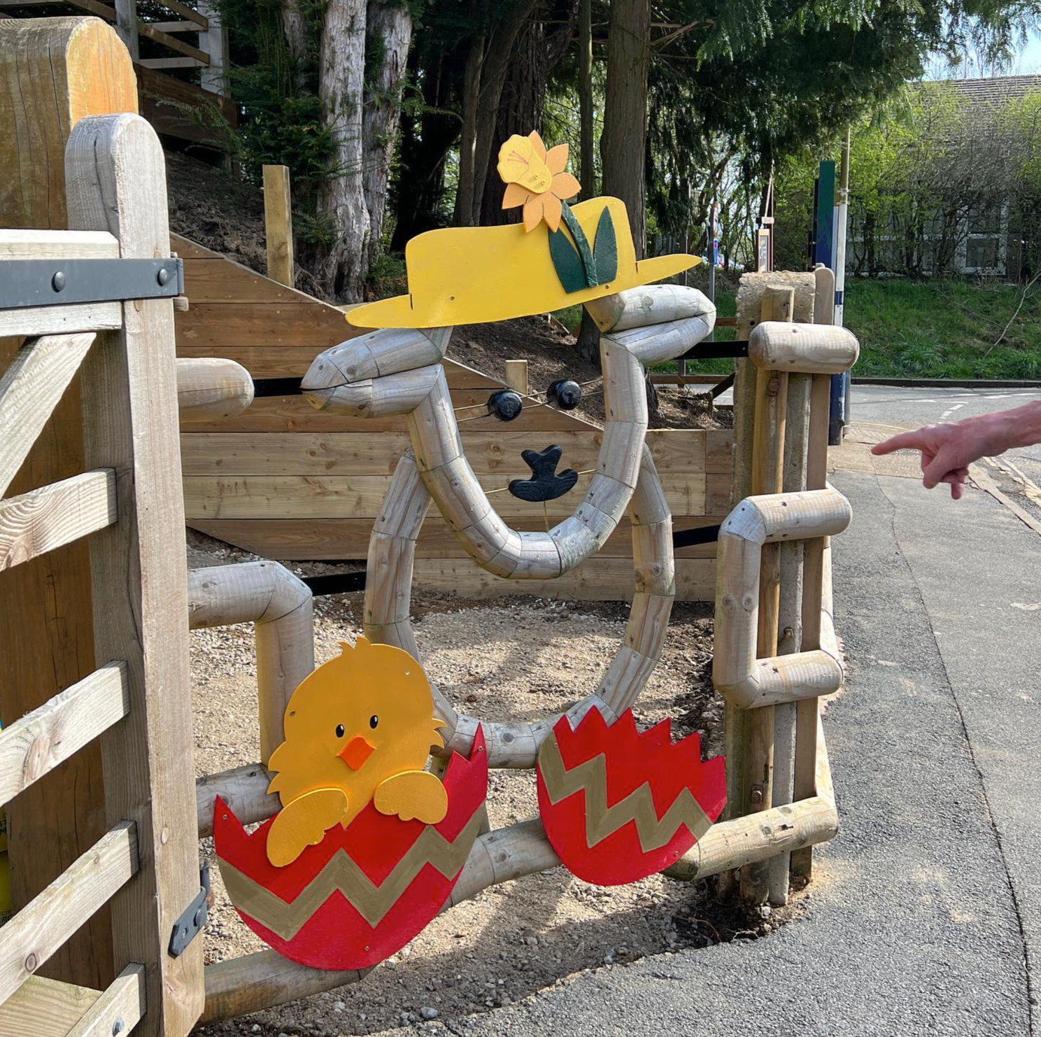 Decorative Easter-themed wooden display with a yellow hat and flower, a chick peeking out of a red egg, and a loop of wooden sticks.