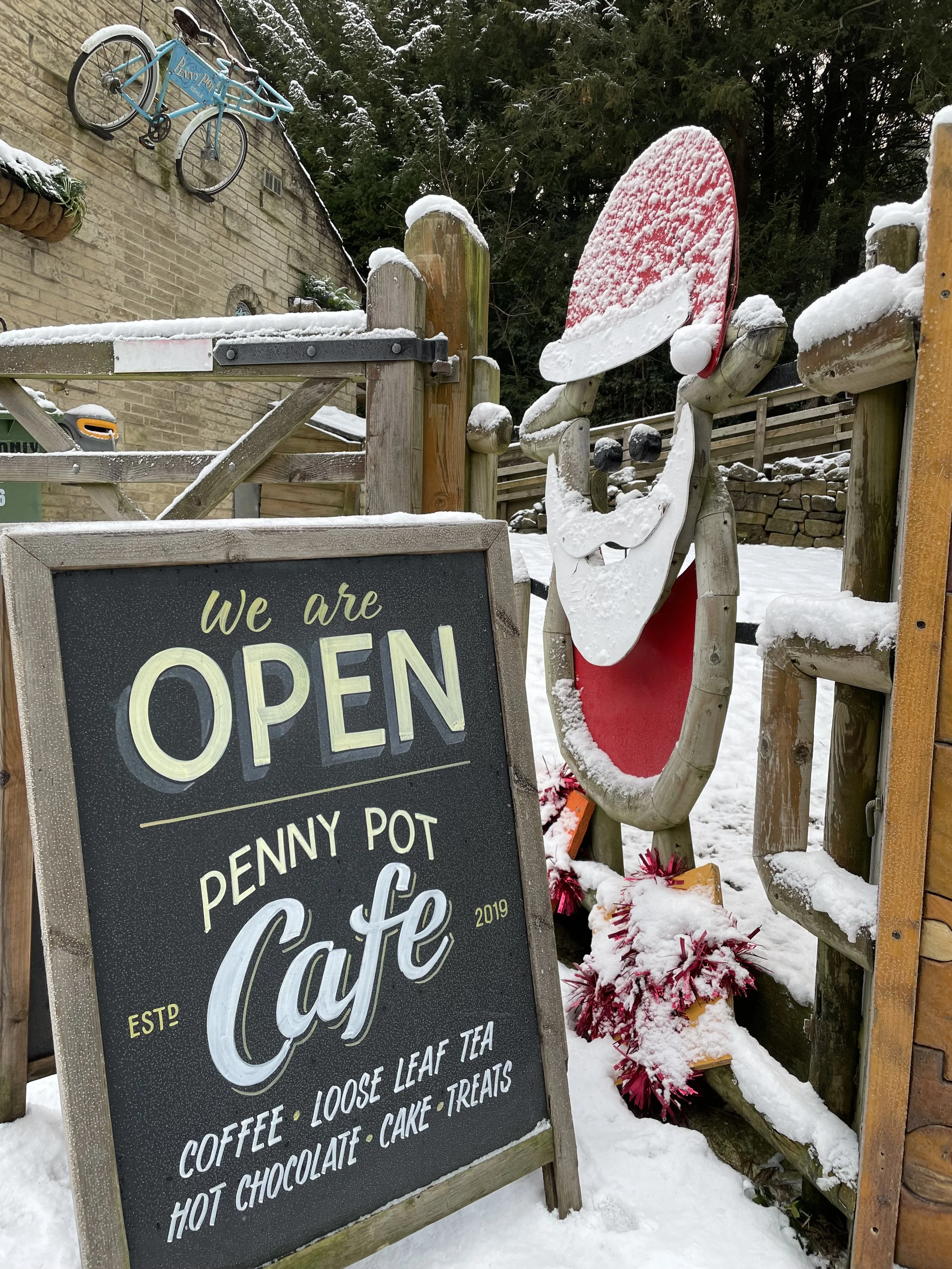 Snow-covered outdoor scene of a cafe with a blackboard sign that reads 'We are open Penny Pot Cafe, Est 2019, Coffee, Loose leaf tea, Hot chocolate, Cake, Treats.' There is a snow-covered painted statue of a gnome with a red hat and beard, and a snow