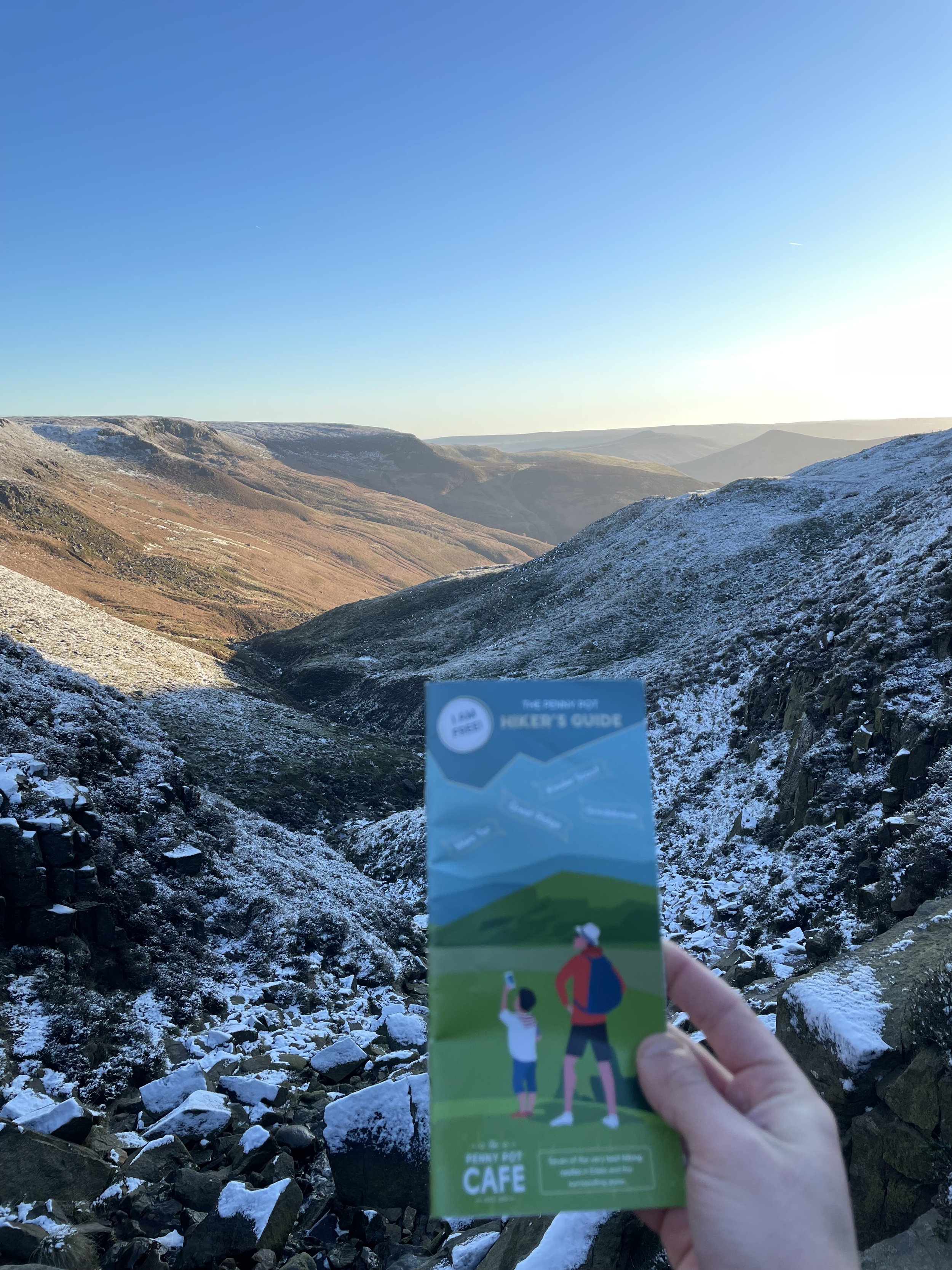 A person holding a colorful hiking trail map with mountains in the background, some snow on the ground, and a clear blue sky.