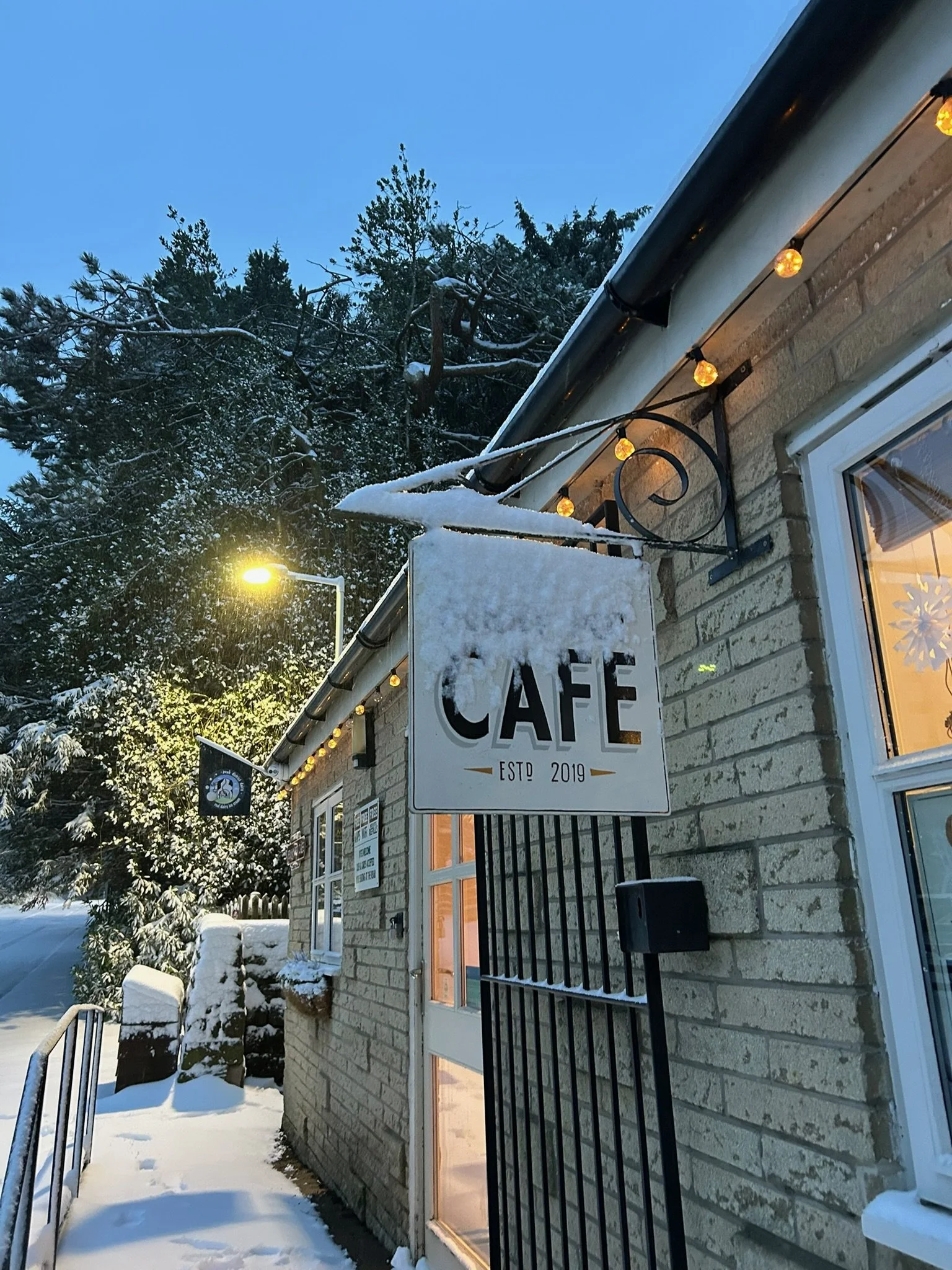 Snow-covered cafe exterior with hanging sign, string lights, and nearby trees.