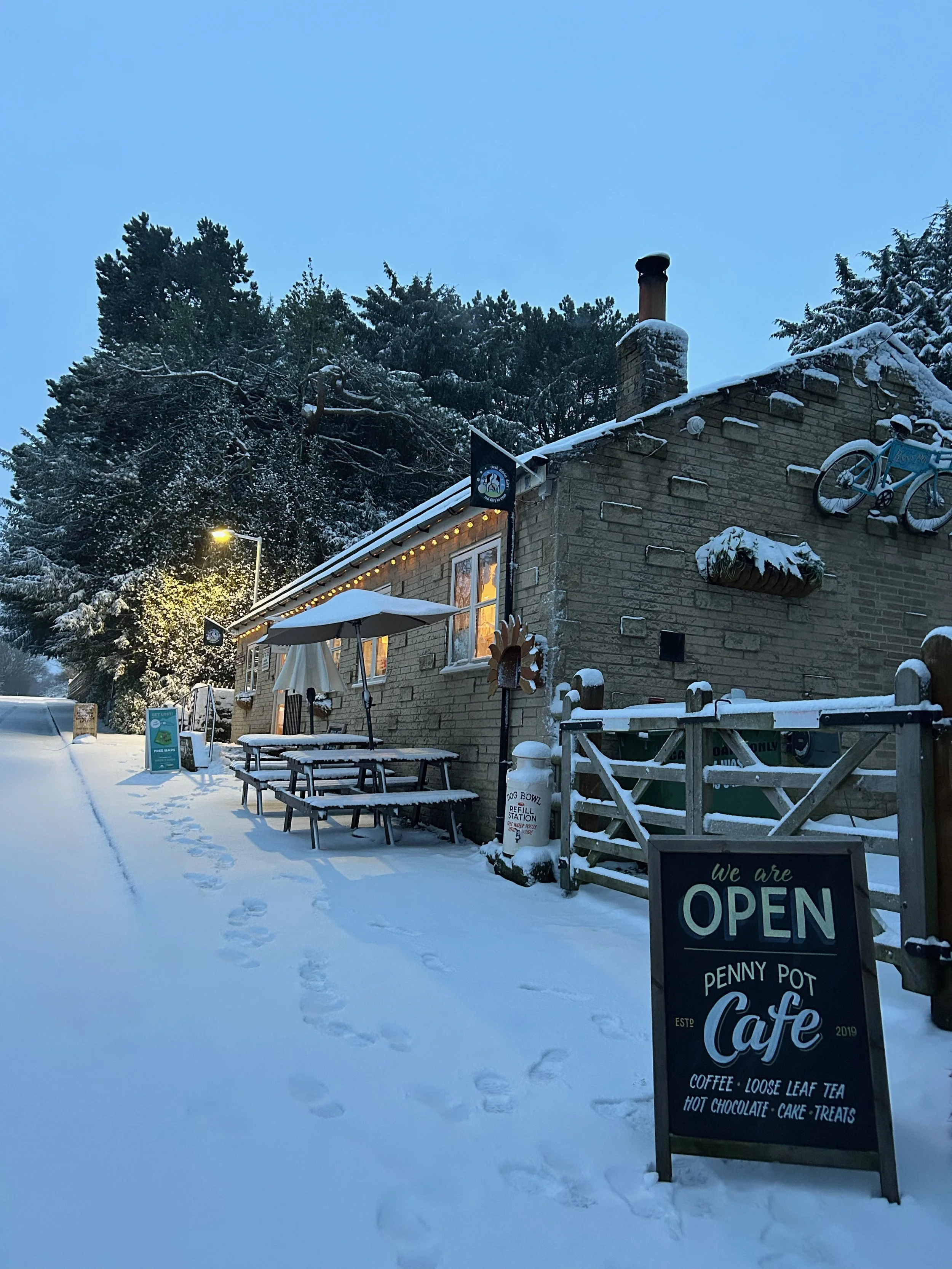 A snowy scene outside a cafe with a sign that reads "We are OPEN Penny Pot Cafe." The cafe is a brick building with outdoor seating, including picnic tables and an umbrella, covered in snow. There is a bicycle mounted on the building's wall and trees