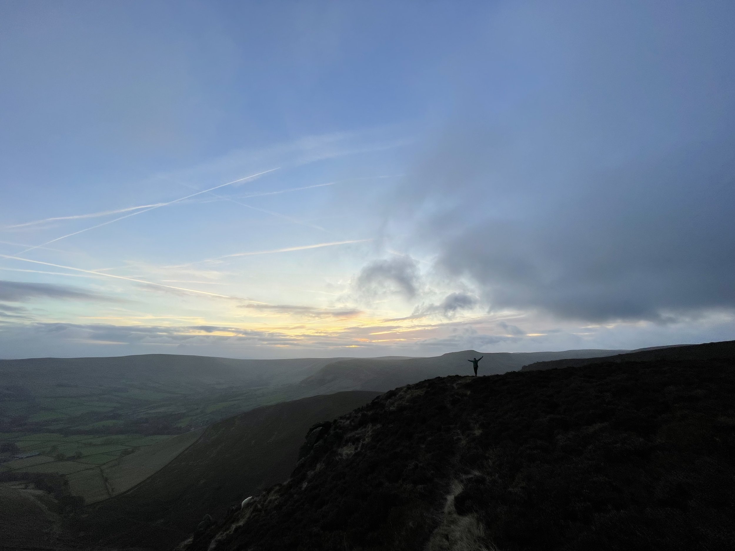 A person standing on a hilltop with arms raised during sunset, overlooking a valley with rolling hills and a partly cloudy sky.