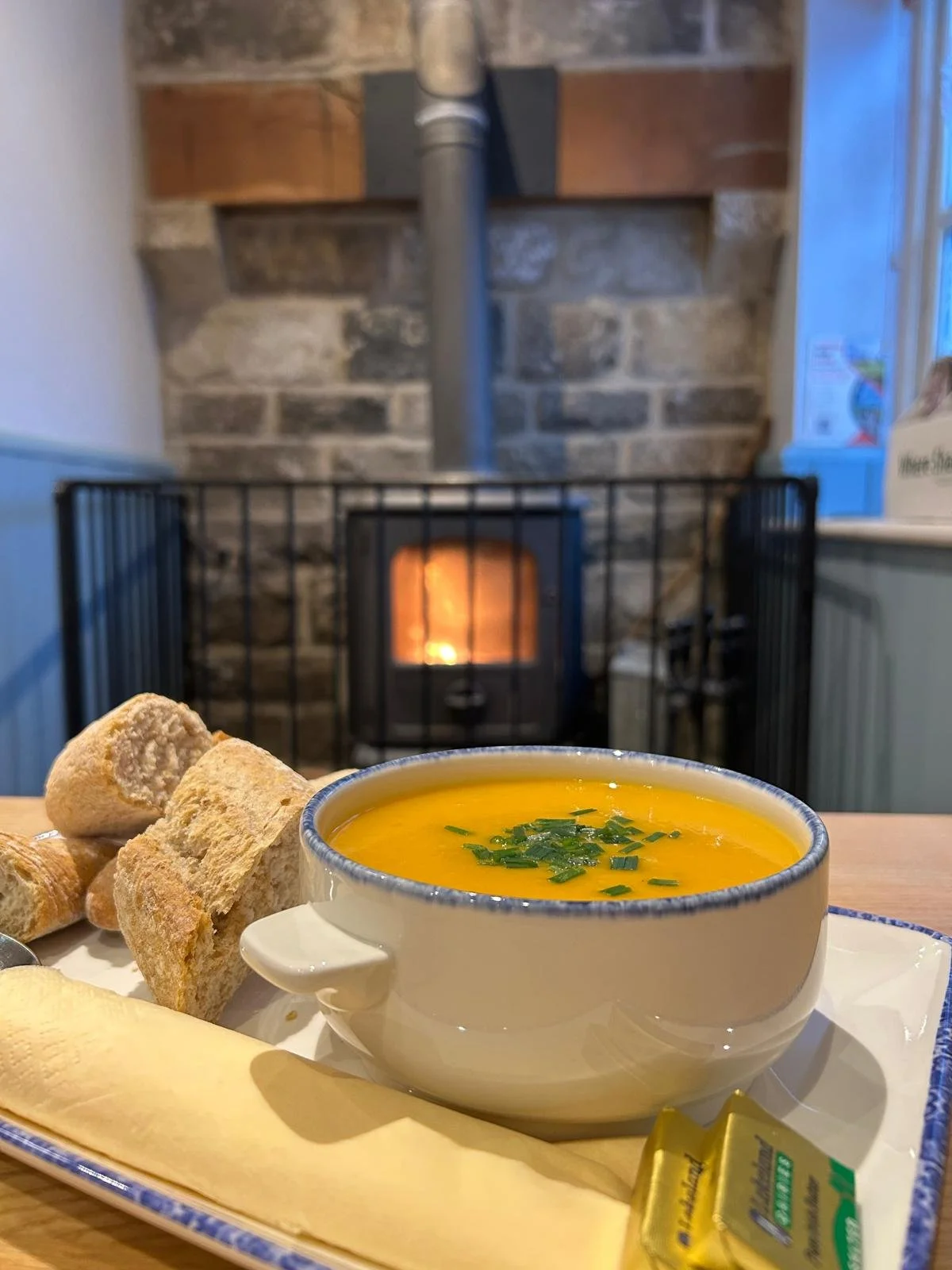 A bowl of yellow soup garnished with green herbs on a tray with bread, a slice of cheese, and butter, with a fireplace in the background.