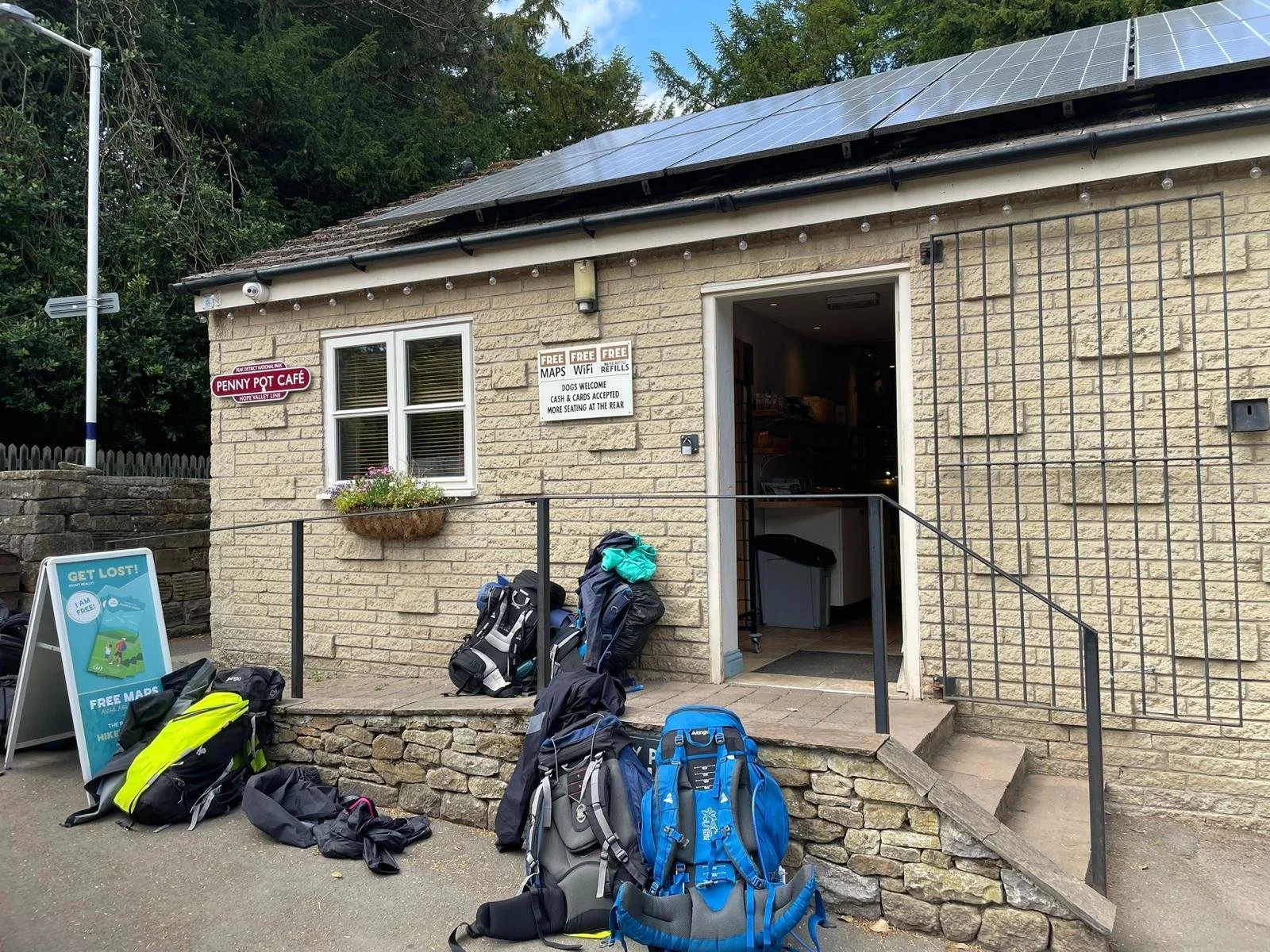 Small brick building with an open door, outdoor backpacks and gear on the ground, signs advertising free maps, WiFi, and dog-friendly policies, and a sign for Penny Pot Cafe.