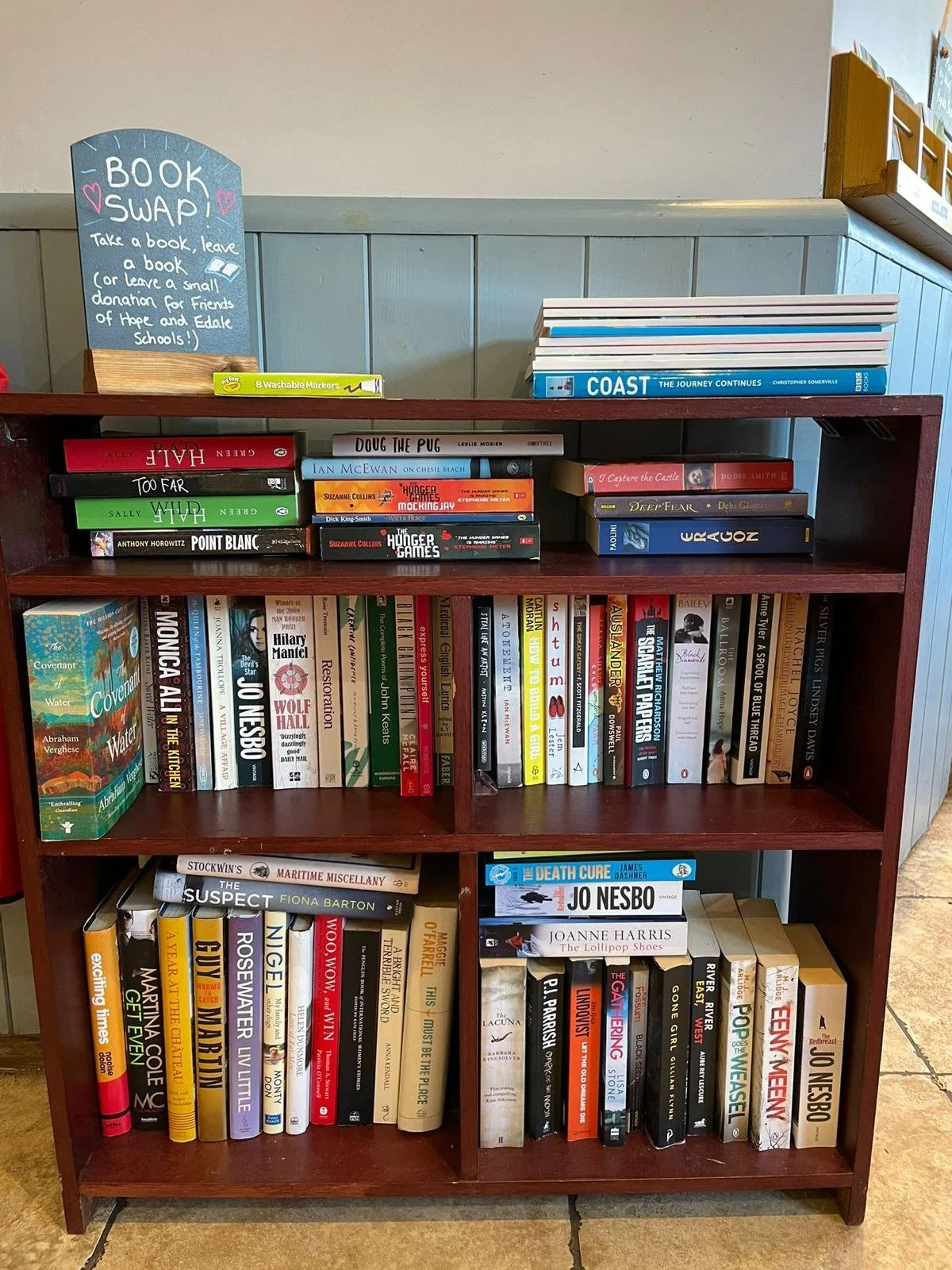 A bookshelf with books organized on three shelves and a small chalkboard sign on top that reads "Book Swap! Take a book, leave a book (or leave a small donation for Friends of Hope and Edale Schools!)."