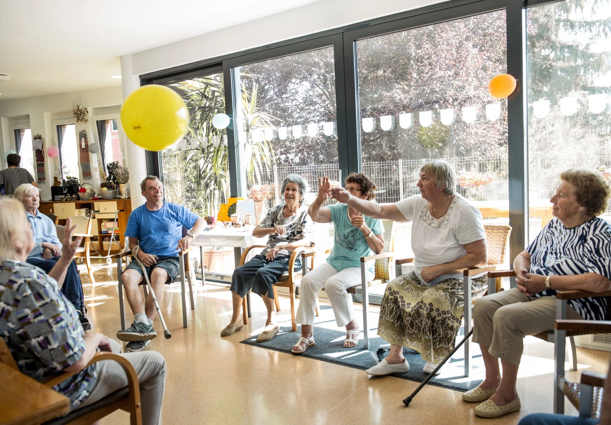 Group of elderly people sitting in chairs in a bright room, playing with colorful balloons during a social gathering.