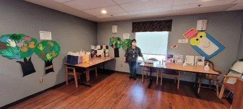 Indoor room with a woman standing near tables with gift bags and a children's artwork wall.