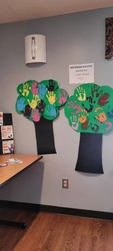 Decorative wall art featuring two trees with colorful handprint leaves, situated in a room with a sign and a framed document on the wall.