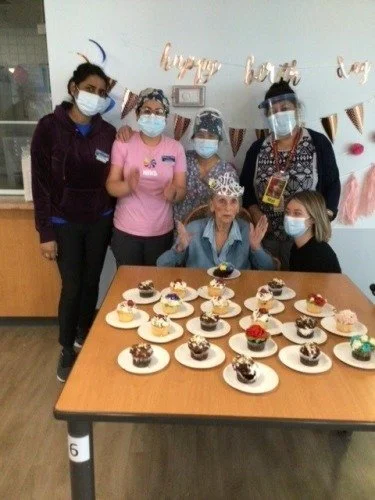 Group of six women, wearing masks and protective face shields, celebrating a birthday with cupcakes on a table.