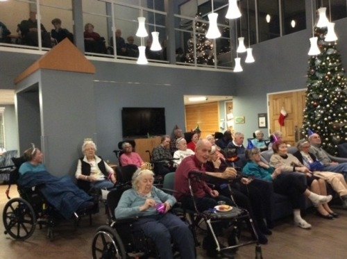 Group of elderly people in a common room with Christmas decorations, some in wheelchairs, watching a performance or event.