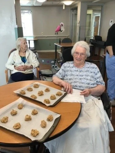 Two elderly women in a care facility, one seated at a table with trays of cookies, the other in a wheelchair nearby.