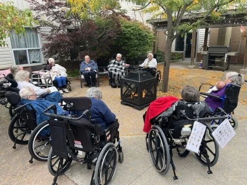 Elderly residents sitting in wheelchairs and chairs outdoors in a courtyard, listening to a group speaker near a small fireplace or fire pit.
