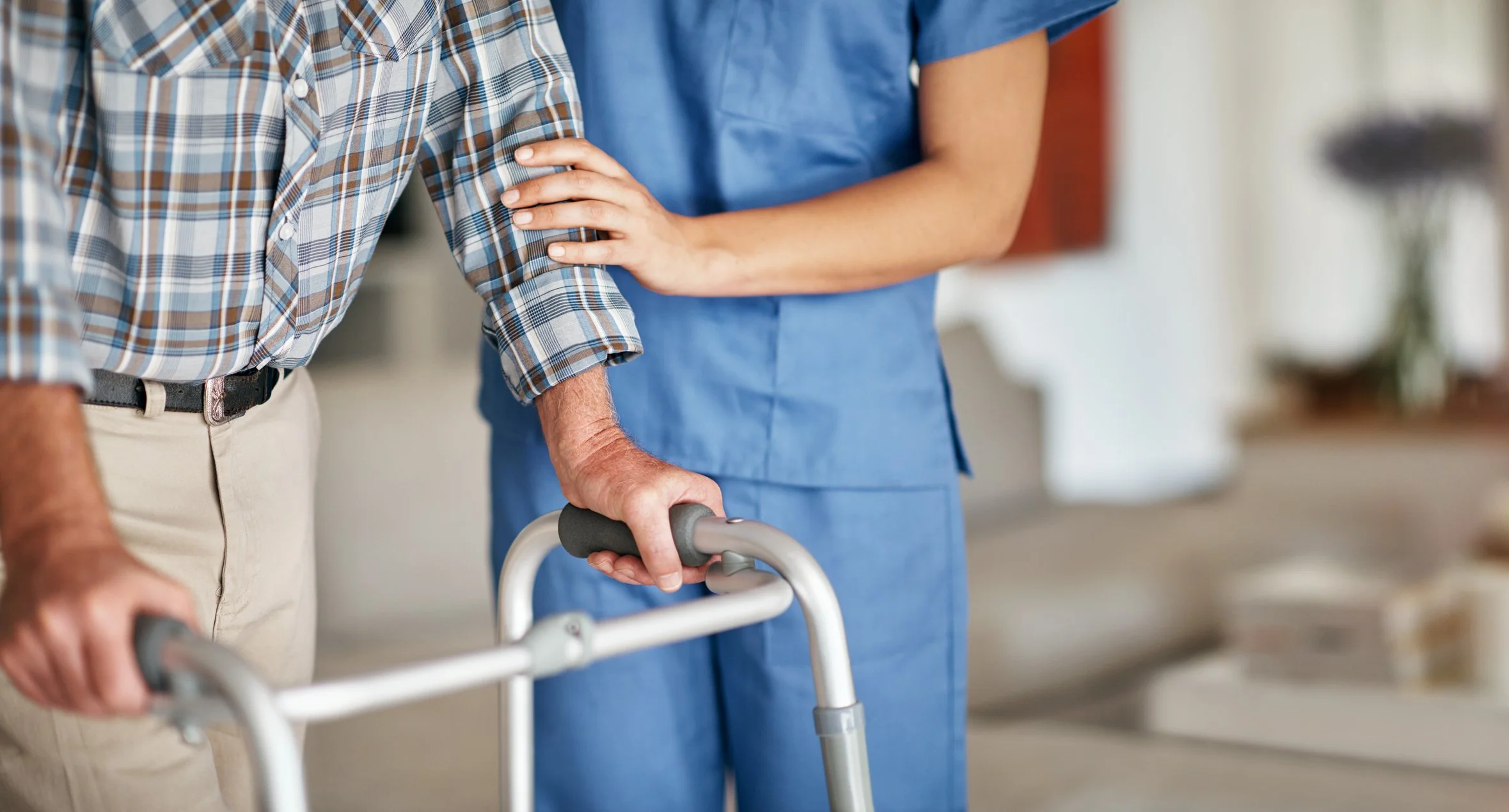 A woman in blue medical scrubs assisting an elderly man with a walker in a home setting.