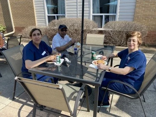 Three people in medical scrubs sitting at a glass outdoor table, eating lunch in a backyard patio area with bushes and a house in the background.