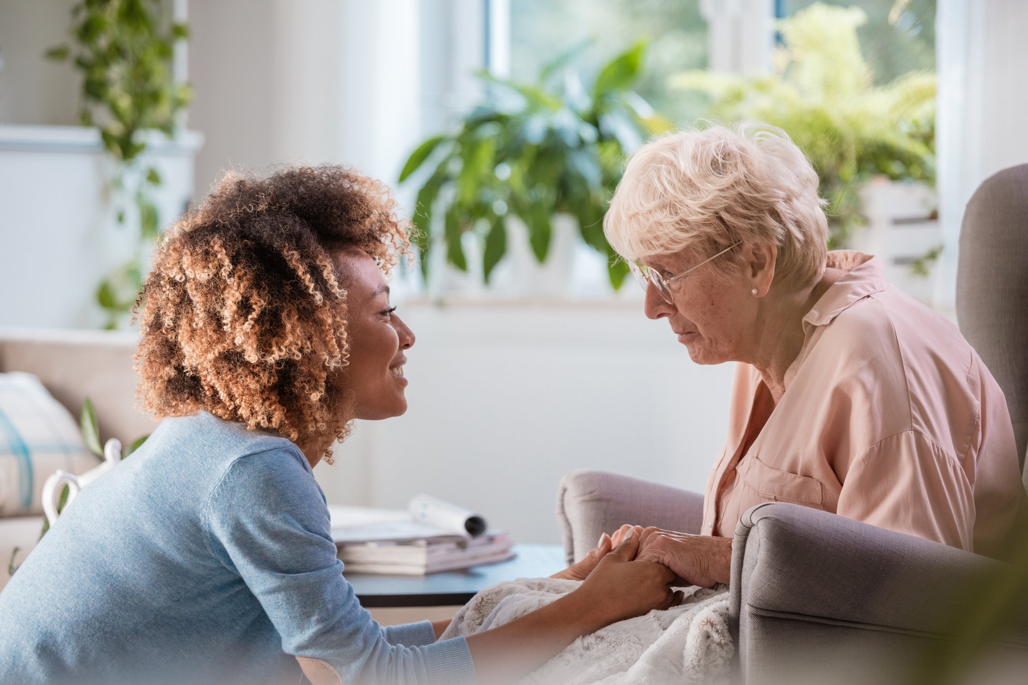 A young woman and an elderly woman share an emotional moment, holding hands and smiling at each other indoors with sunlight and green plants in the background.