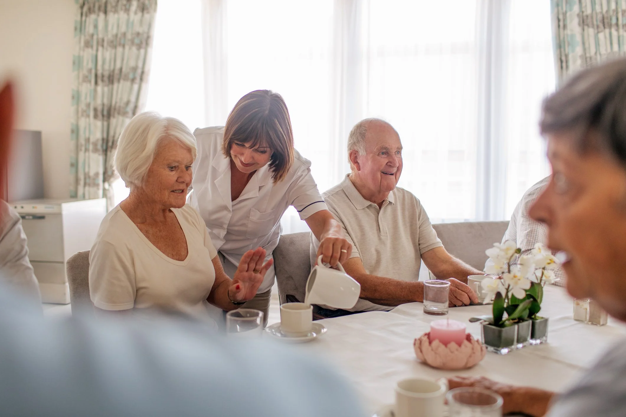 Several elderly people sitting around a dining table during a gathering with a caregiver pouring tea.