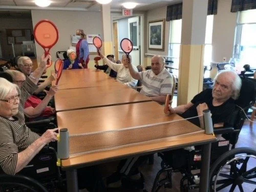 Group of elderly people playing pickleball on a ping-pong table in a recreation room, some in wheelchairs, holding paddles and preparing to rally.