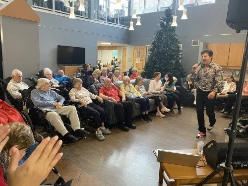 A man standing and speaking to an audience of elderly people seated in a semi-circle in a room decorated with a Christmas tree. The setting appears to be a community or activity center.