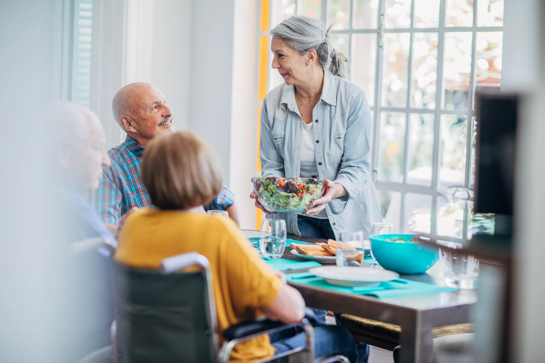 An elderly woman with gray hair serving a salad to a group of seniors at a dining table in a bright room with large windows.