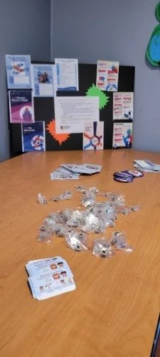 Table with scattered small plastic bags containing buttons, a few stacks of cards, and a black display board with various flyers and informational posters in the background.