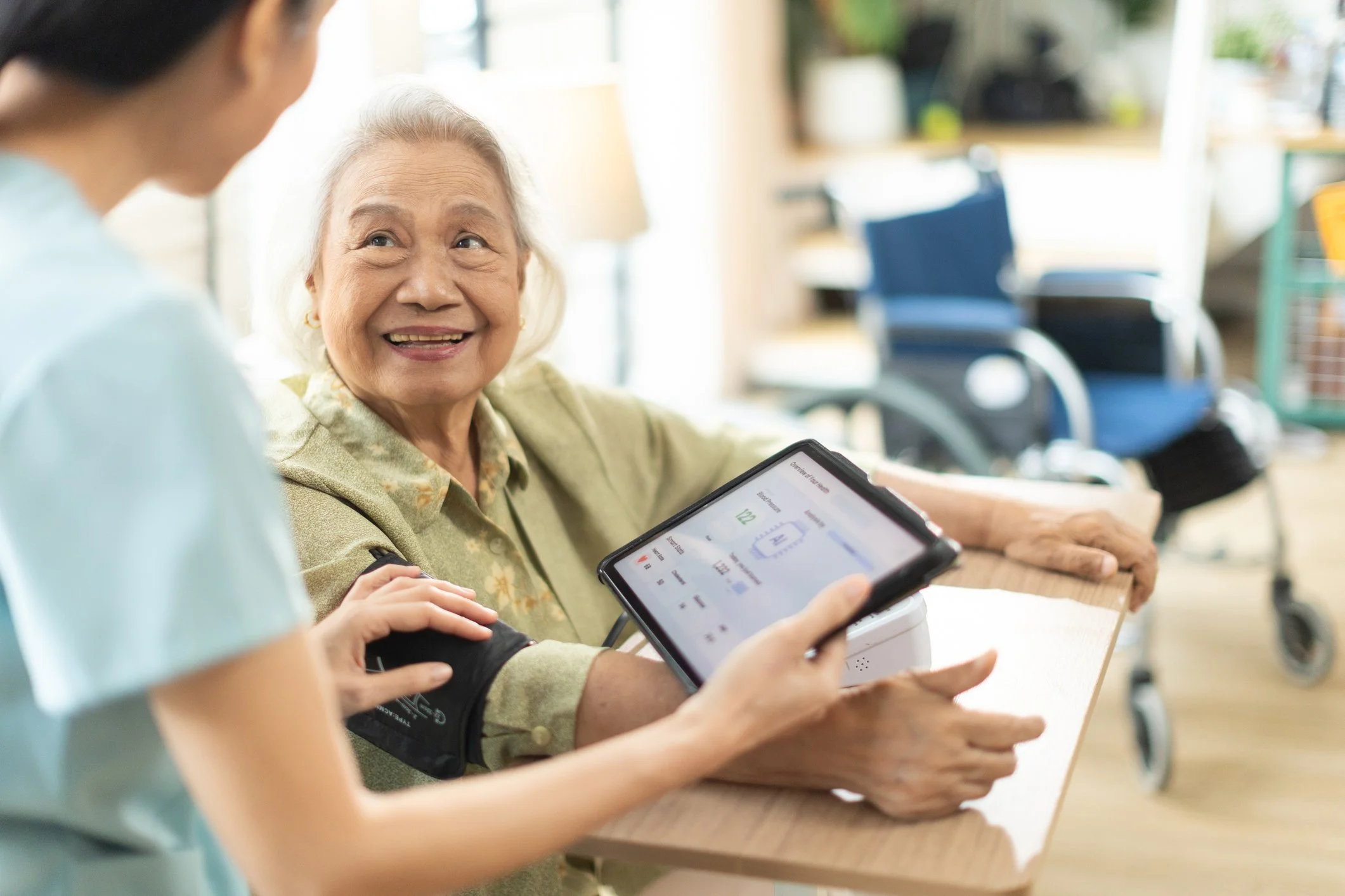 An elderly woman sitting at a table smiling, while a caregiver checks her blood pressure with a medical device. The caregiver holds a tablet displaying health information. There is a wheelchair in the background.