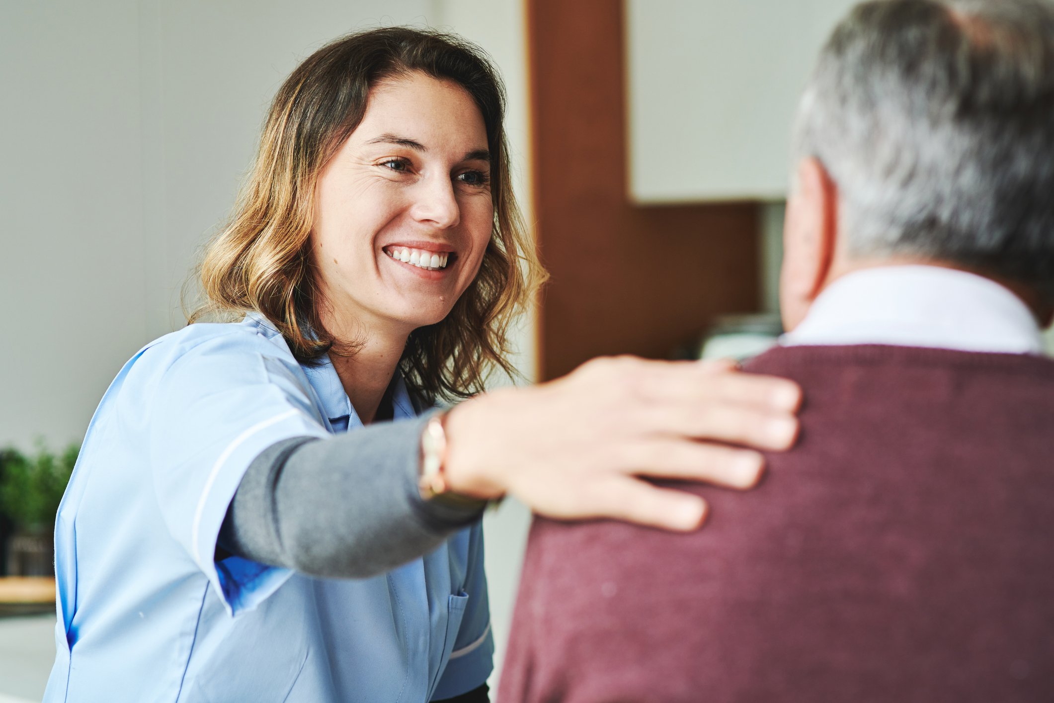 A smiling female nurse gently resting her hand on an elderly patient's shoulder during a consultation in a medical office.