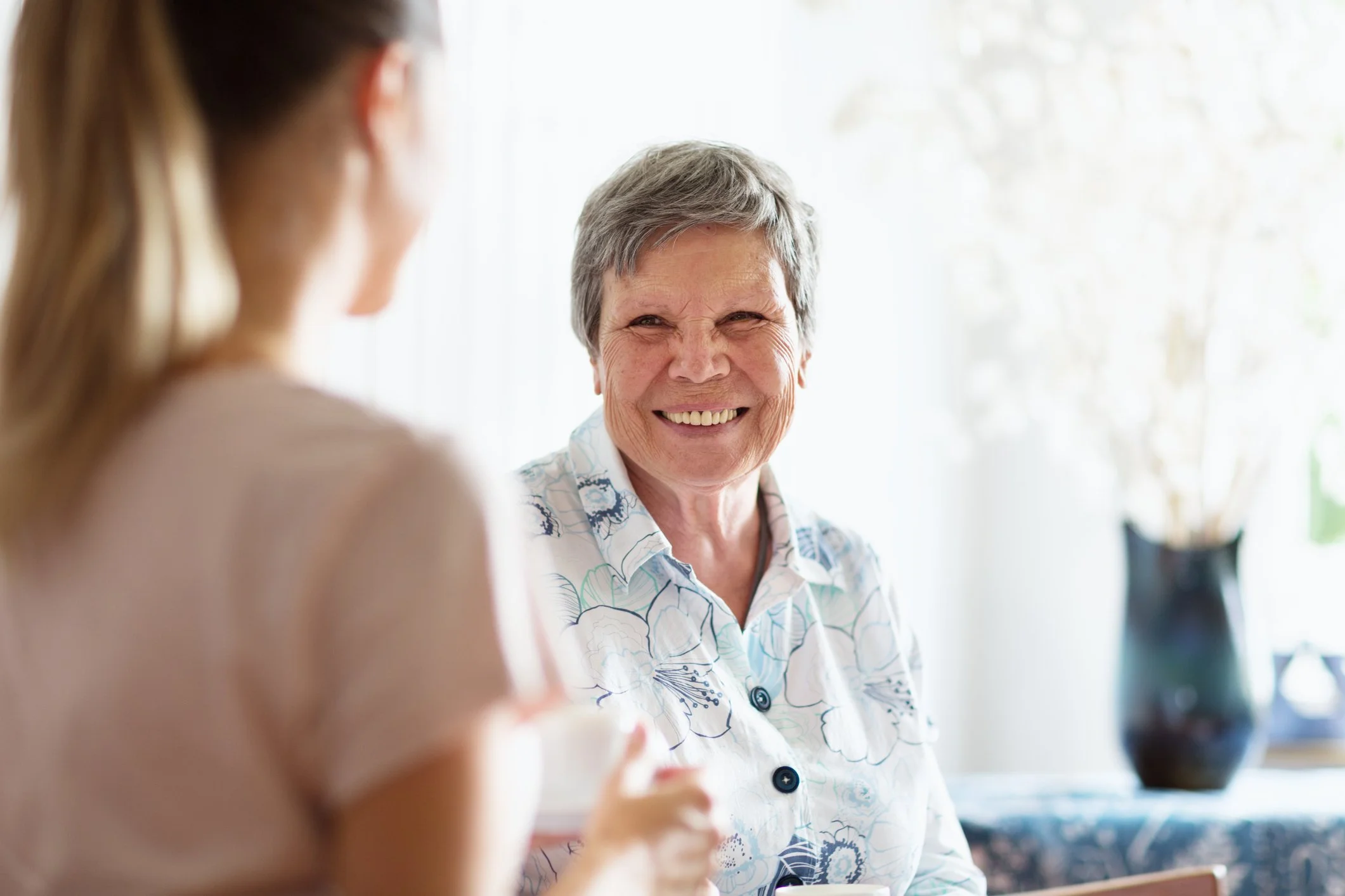 An elderly woman smiling and talking to a younger woman in a bright room with a vase and flowers in the background.