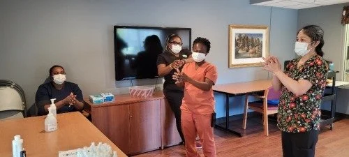 Four women in a room wearing masks, with one woman clapping, two women standing close together, and one woman seated at a table with hand sanitizer on it, in front of a large TV and a framed landscape painting.