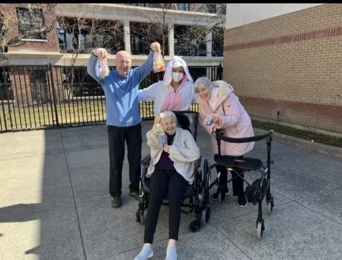 Four elderly women, two in wheelchairs, with two caregivers outside on a paved area near a building, celebrating with smiles and one caregiver holding up small bags.