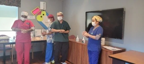 Four healthcare professionals wearing scrubs, masks, and caps standing in a room with a TV and colorful wall decorations.