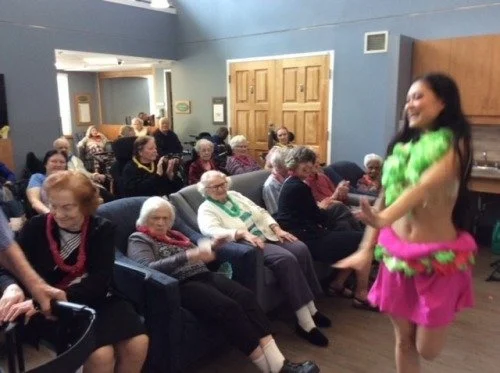 A woman in a colorful costume performing a dance for an audience of elderly people in a community room.