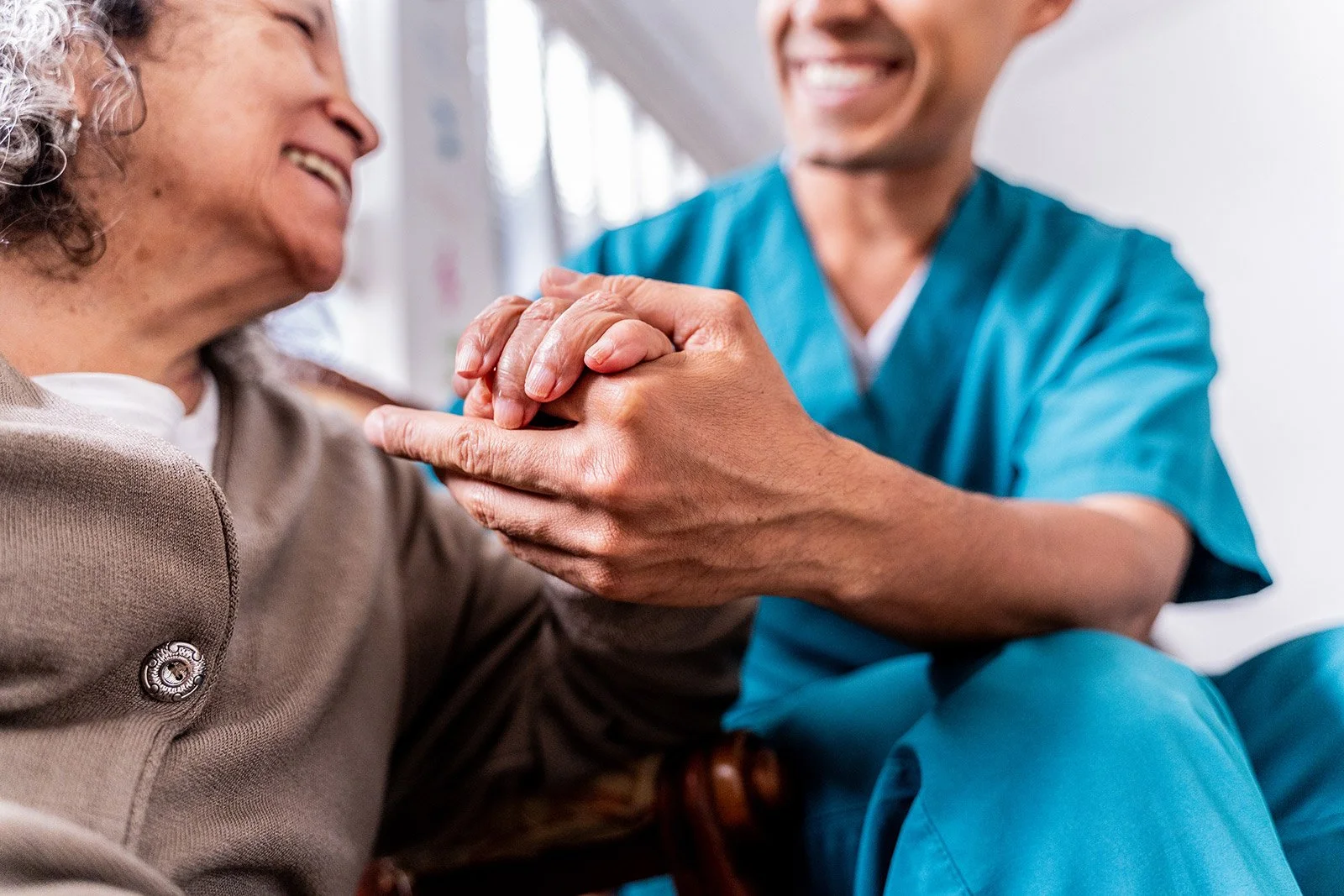 A healthcare professional, dressed in blue scrubs, holding the hands of an elderly woman who is smiling and engaged in a caring moment.