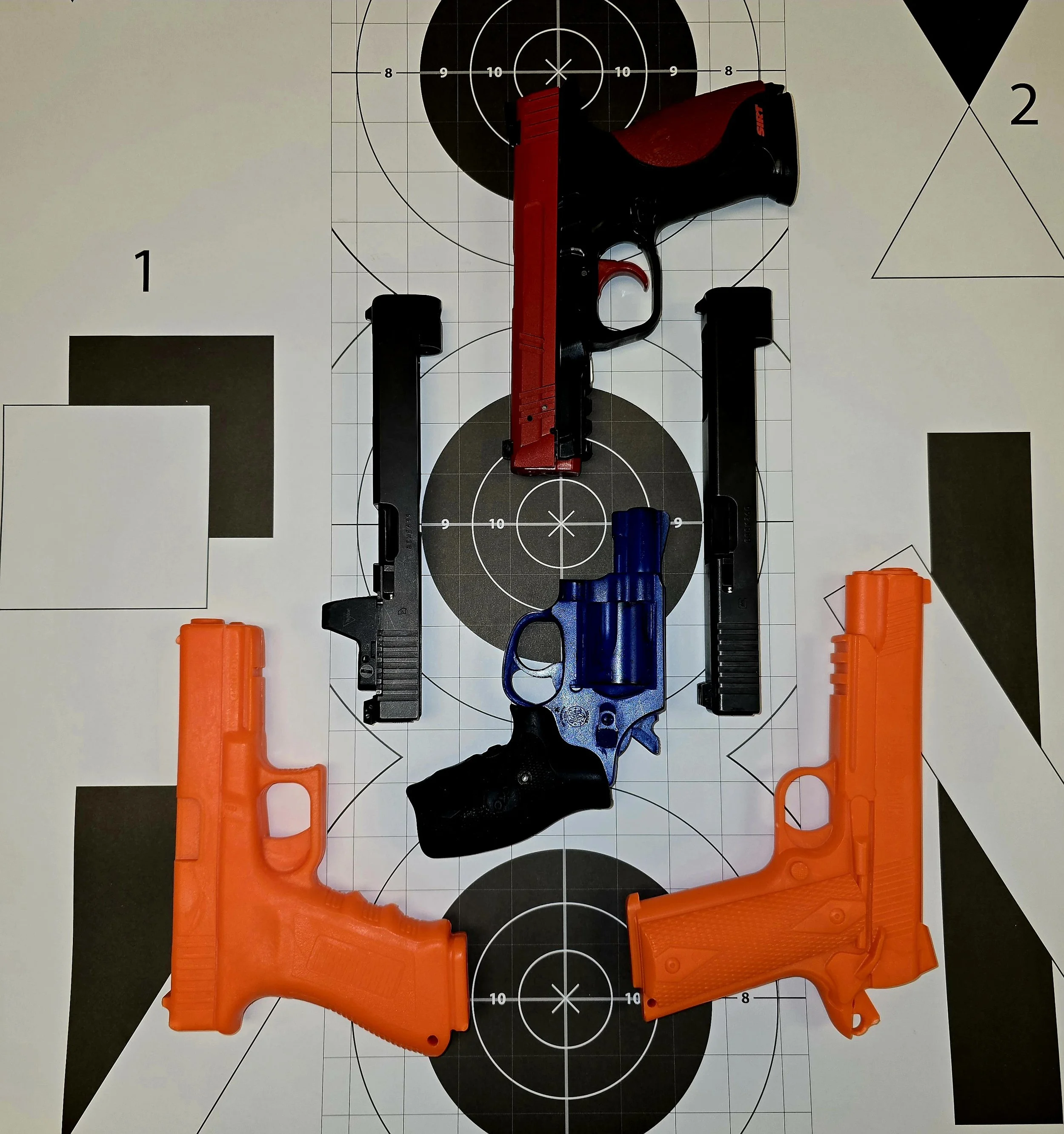 Four simulation guns are arranged on a paper target with multiple black and white circles and geometric shapes, including triangles and squares.