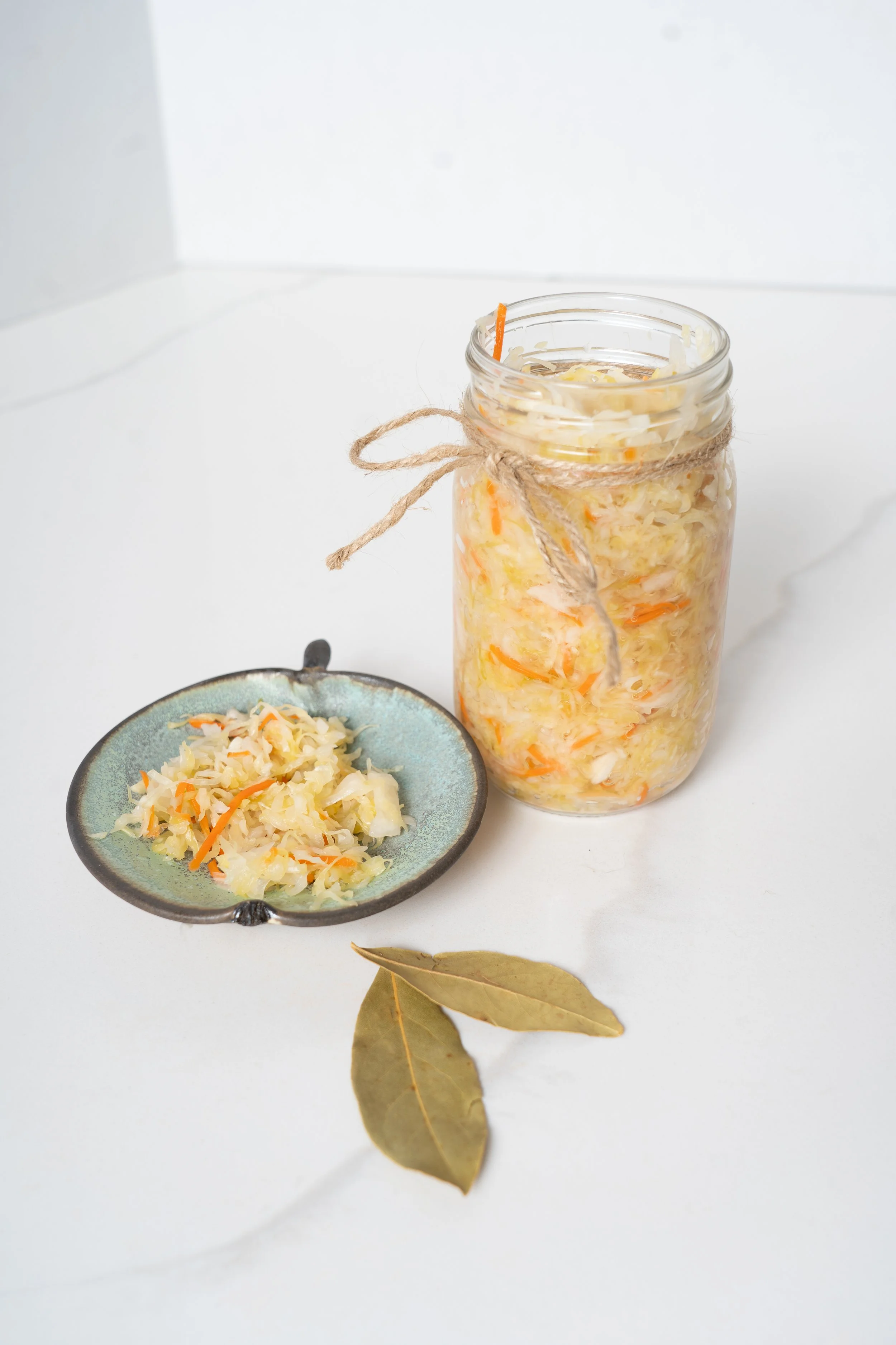 A glass jar filled with sauerkraut, with a piece of twine tied around its neck, placed on a white surface. Next to it is a small blue dish with more sauerkraut, and two dried bay leaves are lying in front.