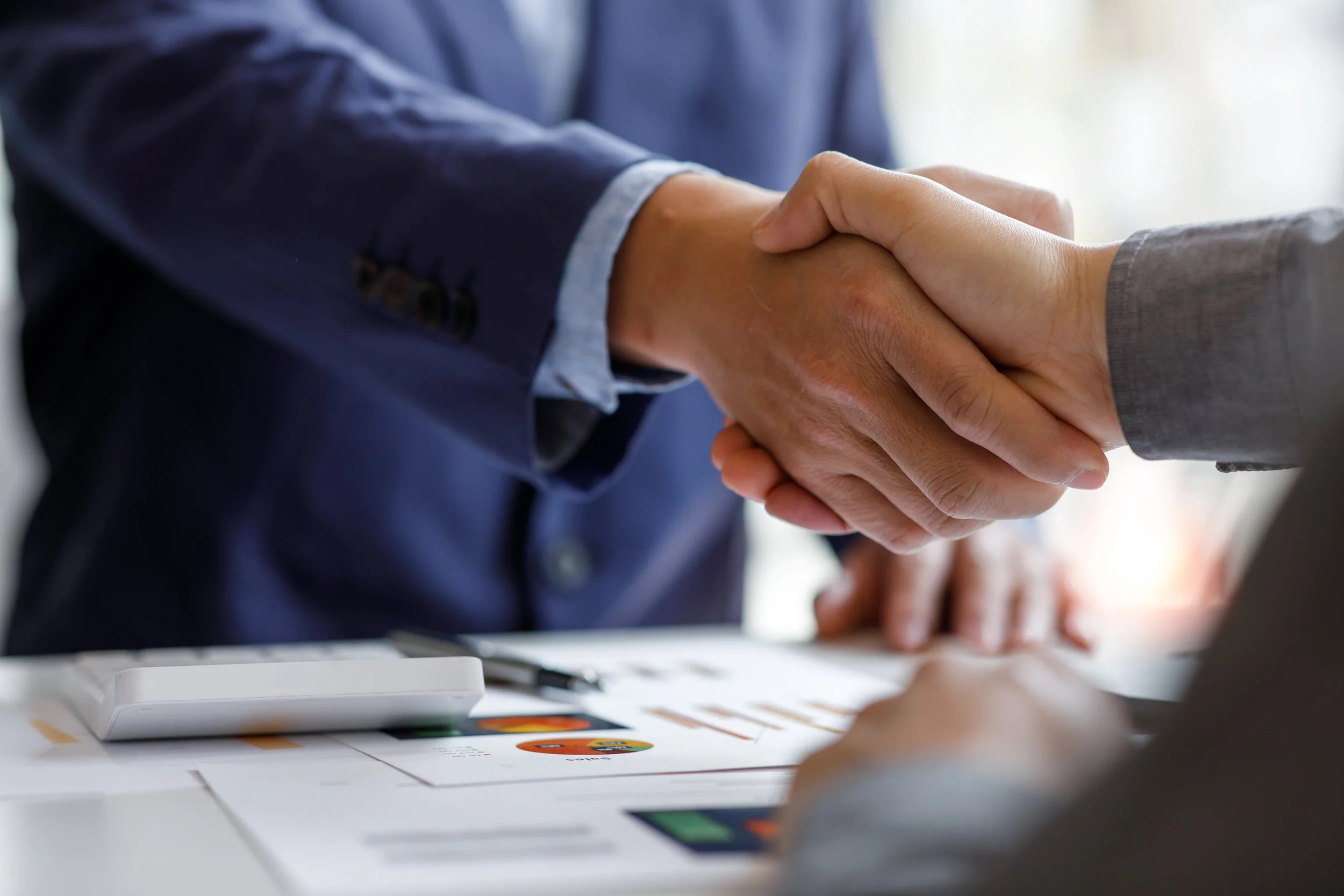 Close-up of two people shaking hands over a table with documents and a calculator, indicating a business agreement or meeting.