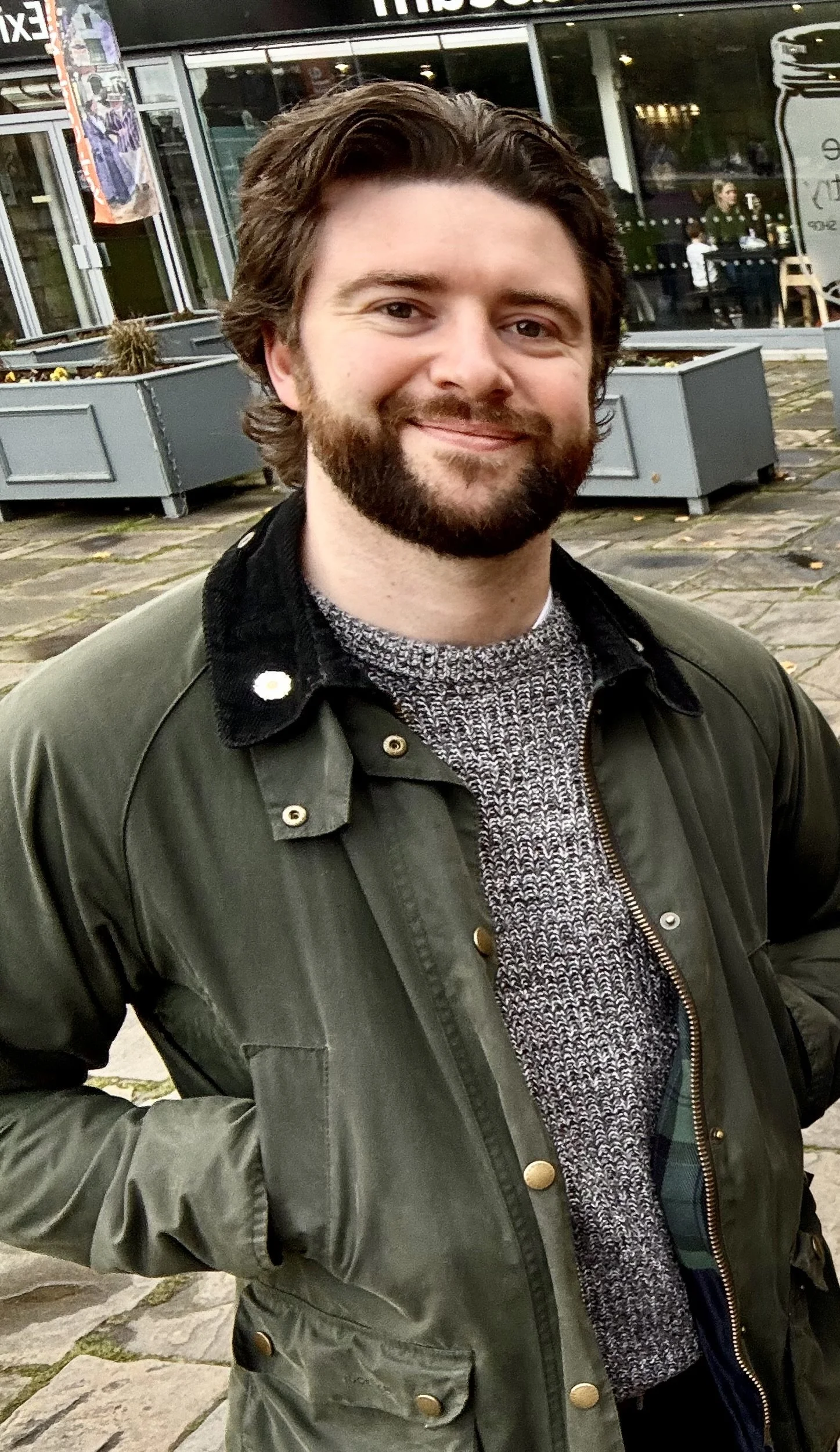 A man with brown hair and a beard smiling outdoors in front of a building with large windows and outdoor seating.