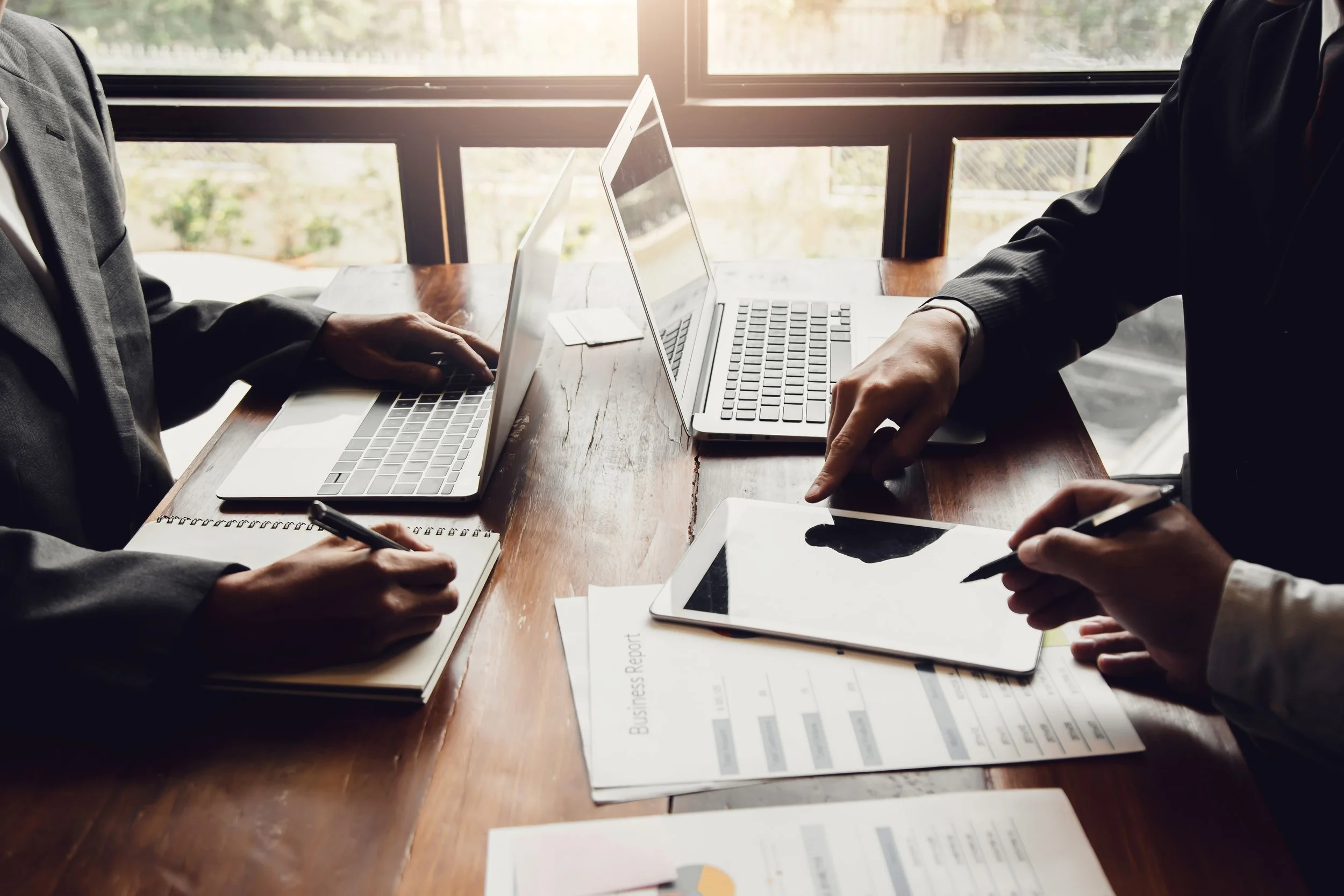 Two people in business suits having a meeting with laptops, tablets, notebooks, and printed reports on a wooden table.