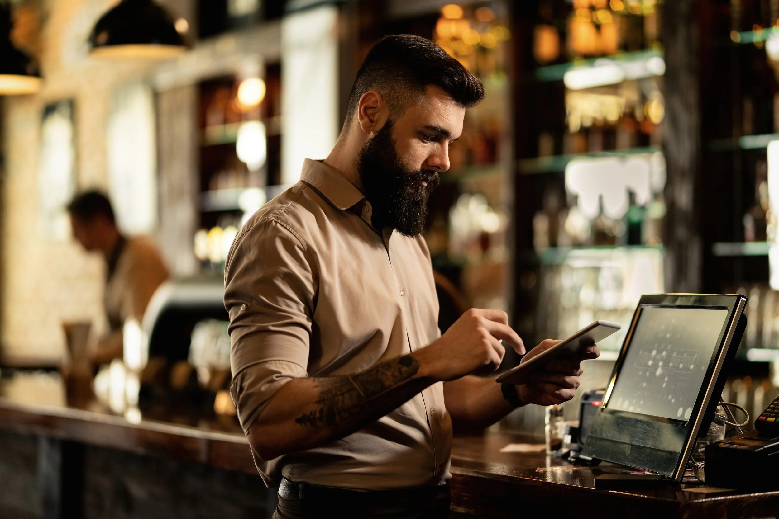 A bearded man with tattoos on his arm, wearing a beige shirt, is standing at a bar counter, using a tablet. The background is blurred, showing shelves with bottles in a warm-lit bar or restaurant.