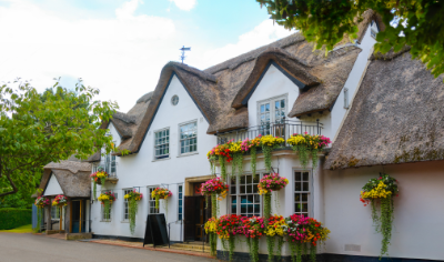 A white building with a thatched roof, decorated with colorful hanging flower baskets and lush green trees, in a charming village scene.