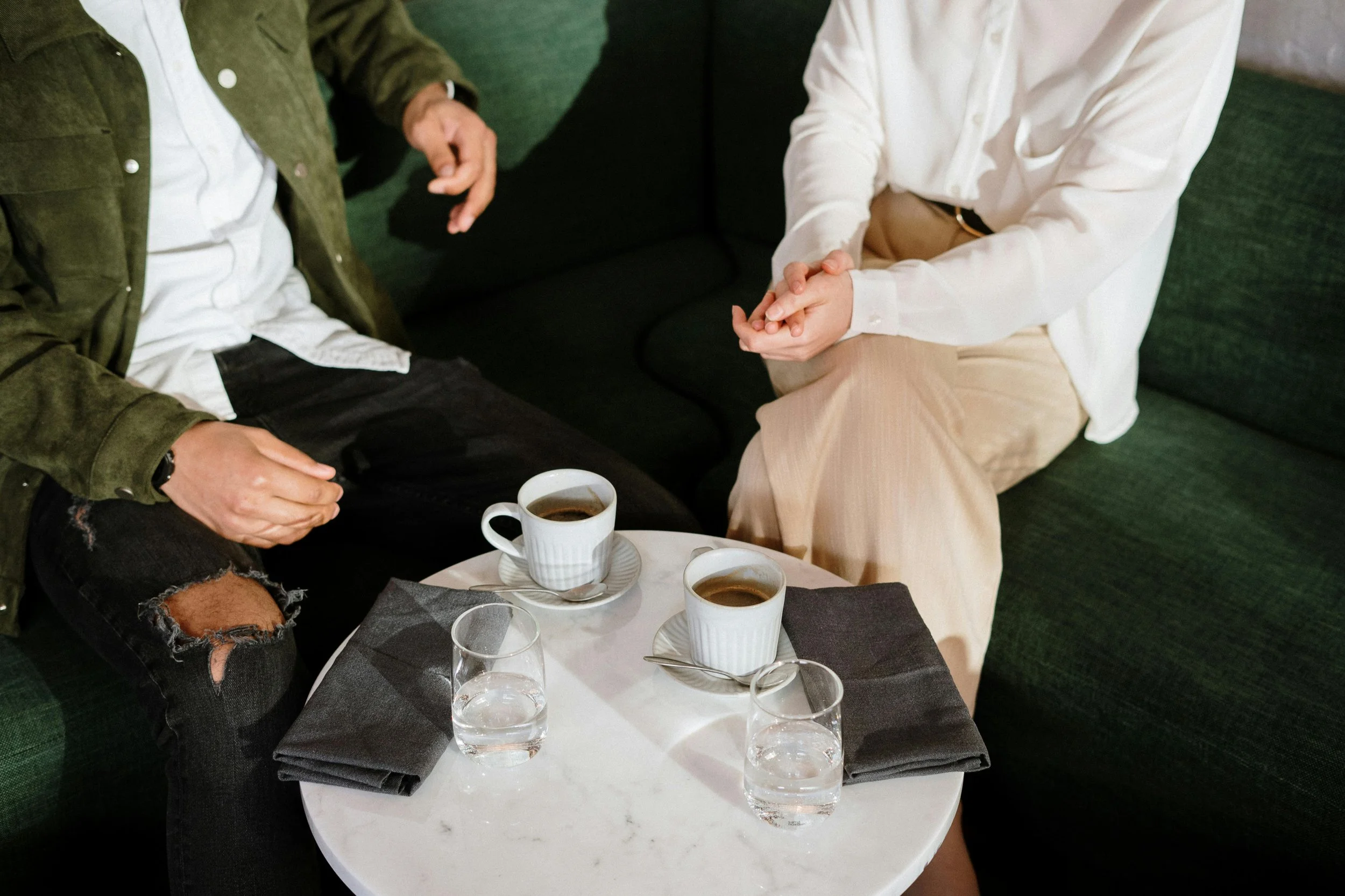 Two people sitting on a green couch having a conversation with a small white table in front of them. The table has two cups of coffee, two glasses of water, and two black napkins.