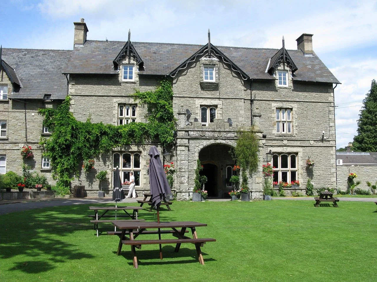 A large stone building with a green lawn and outdoor tables in front. The building has multiple windows and decorative architectural details, with flowers and greenery outside.