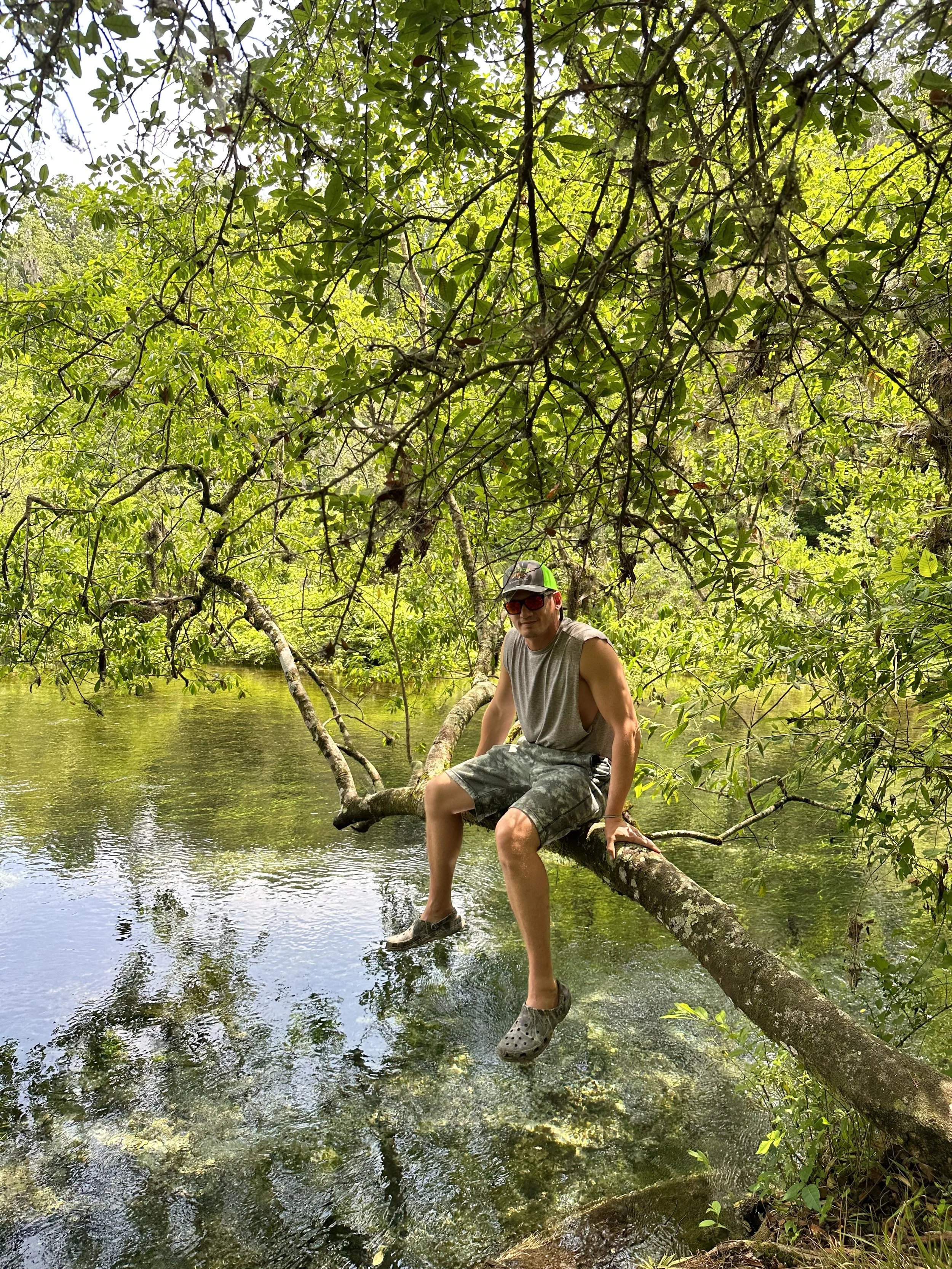 Man sitting on a low-hanging tree branch over a clear river in a lush green forest, wearing a sleeveless shirt, camo shorts, crocs, sunglasses, and a cap.